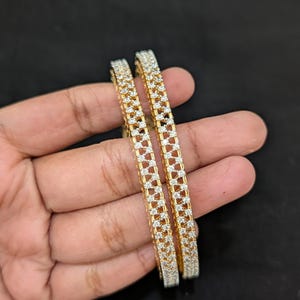 May include: Two gold-toned bangles featuring a lattice-style design encrusted with small, clear, sparkling stones. The bangles have a structured, rectangular profile with a repeating geometric pattern across the outer surface of each piece.
