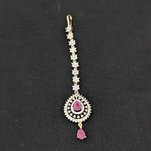 May include: A gold-toned maang tikka featuring a vertical chain of small, clear, star-shaped stones leading to a larger, pear-shaped pendant. The pendant is encrusted with clear stones and centers a single, teardrop-shaped pink stone.