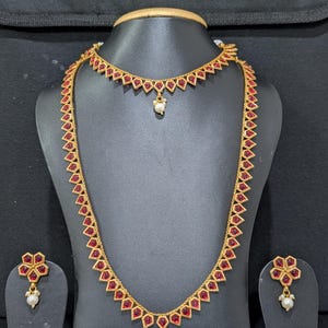 May include: A gold-toned jewelry set featuring a necklace, a choker, and earrings. The set has red gemstone accents and small white pearl drops. The necklace and choker have a geometric design, and the earrings are flower-shaped.