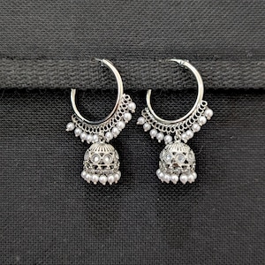 Puede incluir: Un par de pendientes de aro plateados con un colgante en forma de campana jhumka. Los aros están adornados con pequeñas cuentas blancas en el borde inferior, y los colgantes de campana tienen piedras transparentes y una fila de cuentas.