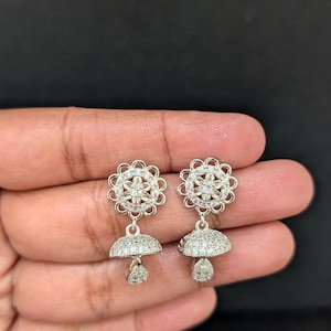 May include: A pair of silver-toned earrings. Each earring features a floral design at the top with a cluster of small, clear stones in the center. Below, a bell-shaped component is also covered in small, clear stones, with a teardrop-shaped stone dangling below.