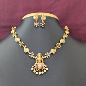 May include: A gold-toned necklace and earring set featuring a central pendant of a deity figure seated on a lotus, accented with small white stones, red gems, and dangling pearls. The chain and earrings display repeating floral and paisley motifs.