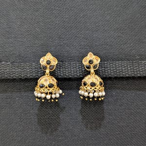 Puede incluir: Un par de pendientes jhumka dorados. Cada pendiente presenta un diseño floral en la parte superior con piedras negras y transparentes. La parte inferior en forma de campana está adornada con piedras negras y pequeñas perlas blancas.