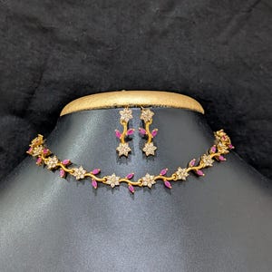 Puede incluir: Un conjunto de collar y pendientes en tono dorado con un diseño de enredadera. La joyería está adornada con pequeñas piedras claras y brillantes dispuestas en forma de flor, intercaladas con piedras de color rosa vibrante.