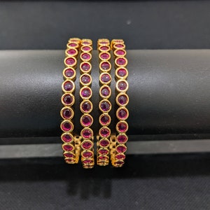 May include: Four gold-colored bangles with a continuous row of round, deep red gemstones. The bangles are displayed on a black surface, showcasing their polished finish and the vibrant contrast between the gold and the red stones.