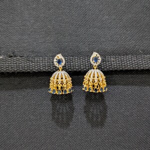 May include: A pair of gold-colored, bell-shaped earrings. Each earring features a filigree design with small, clear stones and blue gemstones. The earrings have a small, teardrop-shaped blue gemstone at the top.