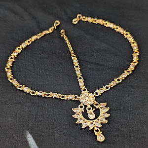 Może przedstawiać: Złoty nakrycie głowy z przezroczystymi, łezkowatymi akcentami. Nakrycie głowy posiada centralny kwiatowy wzór i delikatny łańcuszek, który łączy się z opaską nakrycia głowy. Biżuteria jest prezentowana na czarnej powierzchni.