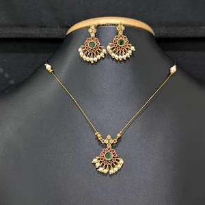 May include: A gold-toned jewelry set featuring a necklace and matching earrings. The necklace has a pendant with a central green stone, surrounded by red stones and small pearls. The earrings mirror the pendant design. The set is displayed on a black mannequin.