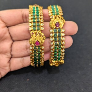 May include: Two gold bangles with emerald green and clear stones. Each bangle features a peacock design with a ruby-colored stone. The bangles are held against a dark background.