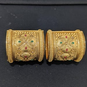 May include: Two gold-colored, ornate bangles with intricate floral and geometric patterns. The bangles feature red and green gemstone accents. The design includes detailed textures and a polished finish, showcasing a traditional jewelry style.