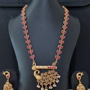 May include: A gold-toned necklace and matching jhumka earring set featuring a central peacock pendant with red stones and white bead accents. The necklace chain consists of repeating leaf-shaped links set with red stones, leading to a flute motif.