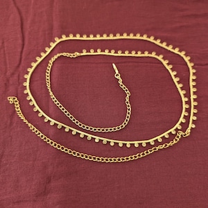 May include: A gold-toned waist chain featuring a delicate linked chain design adorned with small, evenly spaced circular accents. The accessory includes a longer chain segment with a repeating pattern and a shorter, plain curb chain section with a clasp.