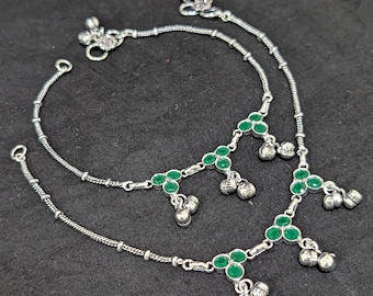 Pode incluir: Duas pulseiras de tornozelo prateadas com detalhes de pedras preciosas verdes e sinos de prata pendurados. As pulseiras de tornozelo são exibidas em uma superfície preta. As pulseiras de tornozelo têm um design de corrente delicado.
