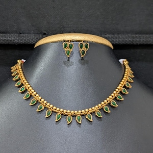 May include: A gold-coloured necklace and matching earrings set. The necklace features a series of green teardrop-shaped stones. The earrings have a cluster of three green stones. The jewellery is displayed on a black mannequin.