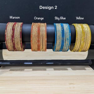 May include: Four sets of bangles labeled Design 2, featuring maroon, orange, sky blue, and yellow colors. Each set consists of multiple thin, textured metallic bangles with a repeating diagonal pattern, displayed on a horizontal black rod.