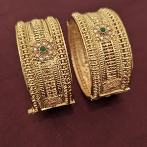 May include: A pair of gold-toned hinged bangles featuring intricate metalwork, textured bands, and floral motifs. Each bangle is accented with a central green stone surrounded by small clear stones and a smaller pink stone at the base.
