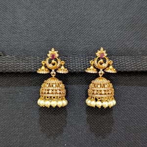 Op de afbeelding: Een paar goudkleurige jhumka-oorbellen. Elke oorbel heeft een klokvormig ontwerp met kleine witte parels die aan de onderkant hangen. De oorbellen zijn versierd met heldere kristallen en een rode edelsteen.