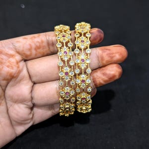 May include: Two gold-colored bangles with intricate floral designs. Each bangle features a series of clear, round gemstones and red, round gemstones. The bangles are displayed against a dark background.