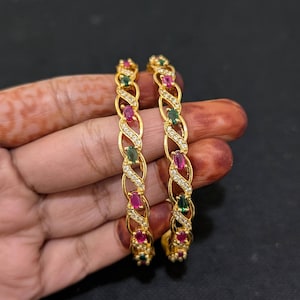 May include: A pair of gold-coloured bangles with a detailed design. Each bangle is adorned with alternating ruby, emerald, and clear gemstone accents. The bangles have a twisted rope-like pattern with small, sparkling stones between the larger gems.