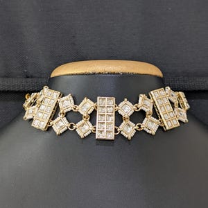 Pode incluir: Um colar tipo gargantilha em tom dourado com uma série de elos retangulares e em forma de diamante. Cada elo é cravejado com pedras transparentes de corte quadrado e contornado por pequenas contas brancas. O design é simétrico.
