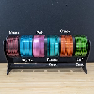 Può includere: Un set di dieci braccialetti di vetro colorati in marrone, azzurro, rosa, verde pavone, arancione e verde foglia. I braccialetti sono disposti su un supporto nero con i nomi dei colori stampati sotto ogni set.