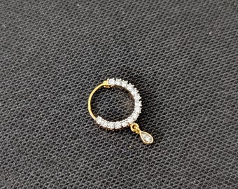 May include: A gold nose ring featuring a circular band encrusted with small clear stones and a single teardrop-shaped pendant hanging from the bottom, which is also set with a clear stone. The metal has a polished yellow gold finish.