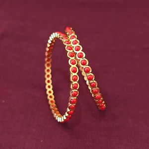 May include: Two circular gold-toned bangles featuring a continuous row of small, round, bright red beads set into the metal frame. The bangles are positioned at an angle, showcasing the metallic gold finish and the vibrant red bead detailing.