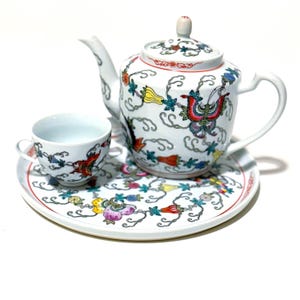 May include: A white porcelain tea set with a teapot, teacup, and serving plate. The set is decorated with colorful floral and butterfly designs. The teapot has a curved spout and handle, and the teacup has a small handle. The plate is round.