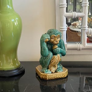 May include: A ceramic monkey figurine in shades of teal, brown, and gold. The monkey is seated, with one hand to its head and the other holding a fruit. It sits on a square, woven-patterned base. A green vase is on the left.