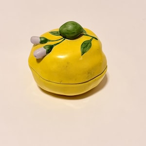 May include: A yellow ceramic trinket box shaped like a lemon, with a green stem and leaves, and two small white flower buds. The box is round and has a lid that fits snugly on top.