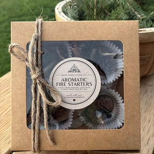 May include: A brown cardboard box tied with twine, containing aromatic fire starters. The box has a clear window showing pine cones and cinnamon sticks. The label reads "AROMATIC FIRE STARTERS".