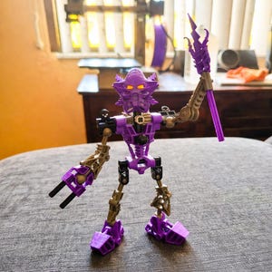 May include: A purple and gold action figure with a detailed design. The figure has a mask-like head with glowing yellow eyes, and is holding a purple staff. The figure is standing on a gray surface.