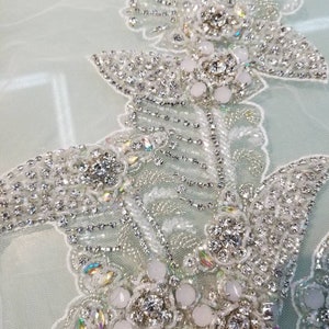 May include: Close-up of a delicate, white beaded lace applique with iridescent rhinestones and pearl-like beads. The applique features a floral design with intricate details.