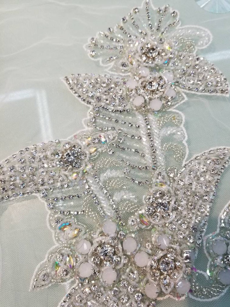 May include: Close-up of a delicate, white beaded lace applique with iridescent rhinestones and pearl-like beads. The applique features a floral design with intricate details.