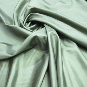 Puede incluir: Una tela suave y sedosa de color verde salvia apagado. La tela está drapeada y doblada, mostrando su textura y brillo.