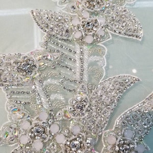 May include: Close-up of a delicate white lace fabric with intricate beading and crystal embellishments. The design features a floral motif with a cluster of white pearls and sparkling rhinestones.