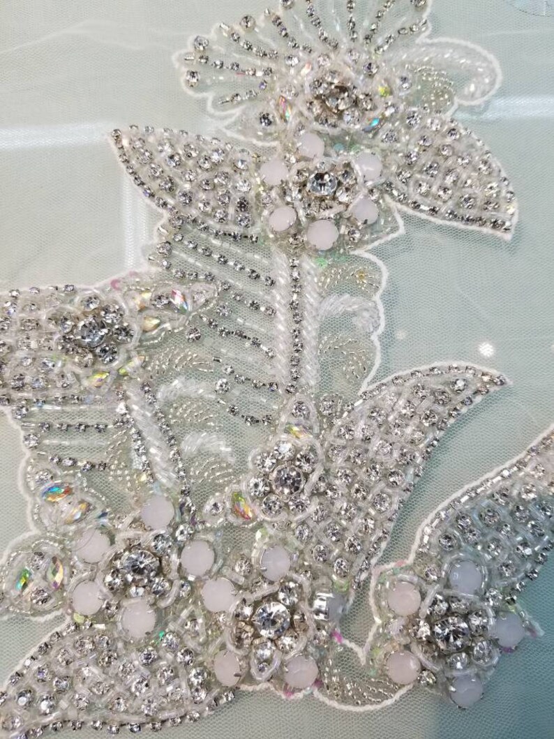 May include: Close-up of a delicate white lace fabric with intricate beading and crystal embellishments. The design features a floral motif with a cluster of white pearls and sparkling rhinestones.