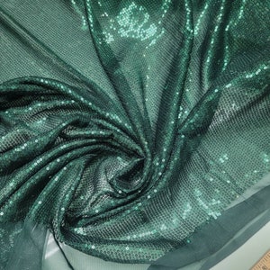May include: Green sequined fabric with a shimmery finish. The fabric is draped and folded, showing the texture of the sequins.