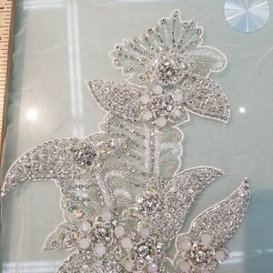 May include: A silver and white beaded floral applique with a delicate design. The applique features a large central flower with smaller flowers and leaves surrounding it. The applique is perfect for adding a touch of elegance to any project.