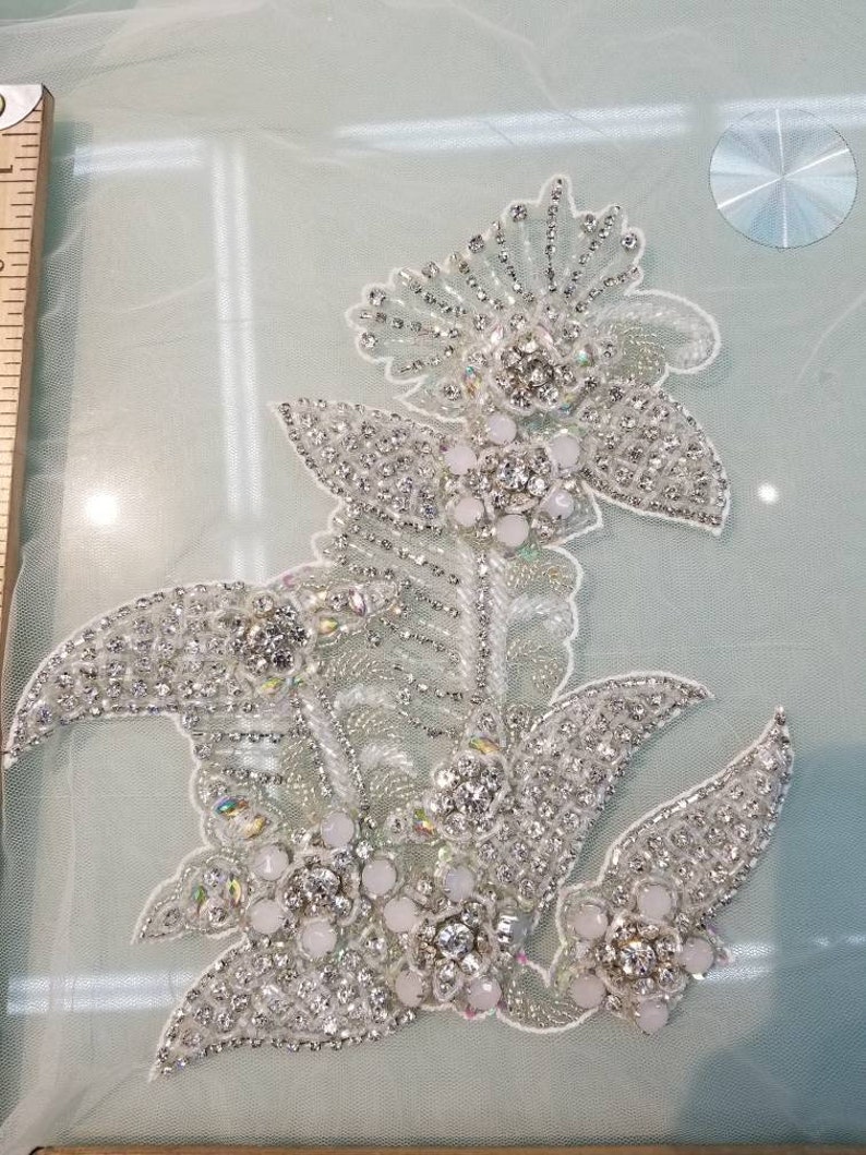 May include: A silver and white beaded floral applique with a delicate design. The applique features a large central flower with smaller flowers and leaves surrounding it. The applique is perfect for adding a touch of elegance to any project.