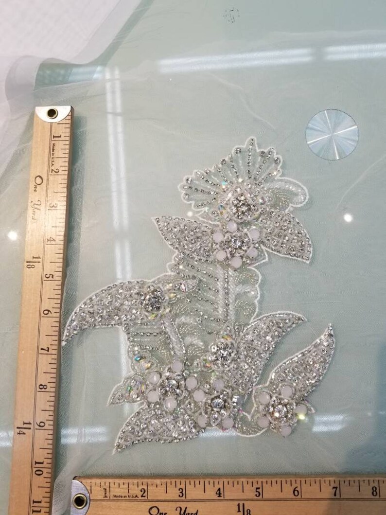 May include: A silver floral applique with white beads and rhinestones. The applique is approximately 5 inches wide and 6 inches tall.