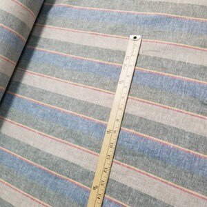 Pu&ograve; includere: Un primo piano di un tessuto a righe con righe blu, rosa e beige. Un righello di legno &egrave; posizionato sopra il tessuto, misurando 43 cm.