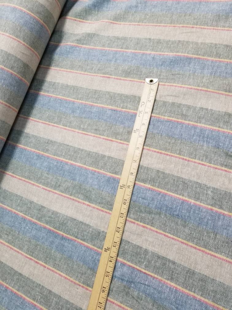 Pu&ograve; includere: Un primo piano di un tessuto a righe con righe blu, rosa e beige. Un righello di legno &egrave; posizionato sopra il tessuto, misurando 43 cm.