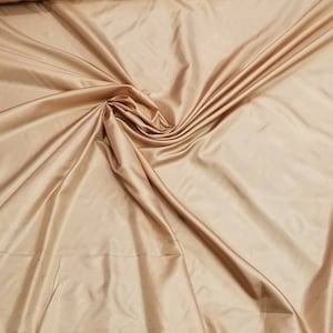 May include: A close-up shot of a large piece of fabric. The fabric is a light tan color with a satin finish. The fabric is draped and folded, creating a rippled effect with highlights and shadows.