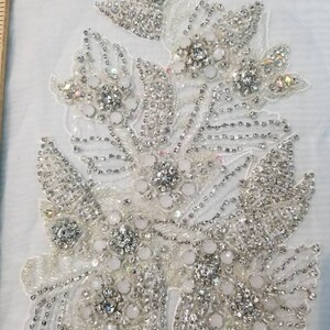 Peut inclure: Une applique florale blanche avec des perles et des strass &eacute;tincelants. Le motif pr&eacute;sente des grappes de fleurs et de feuilles, avec un m&eacute;lange d'ornements clairs et iris&eacute;s. L'applique est probablement destin&eacute;e &agrave; orner des v&ecirc;tements ou des accessoires.