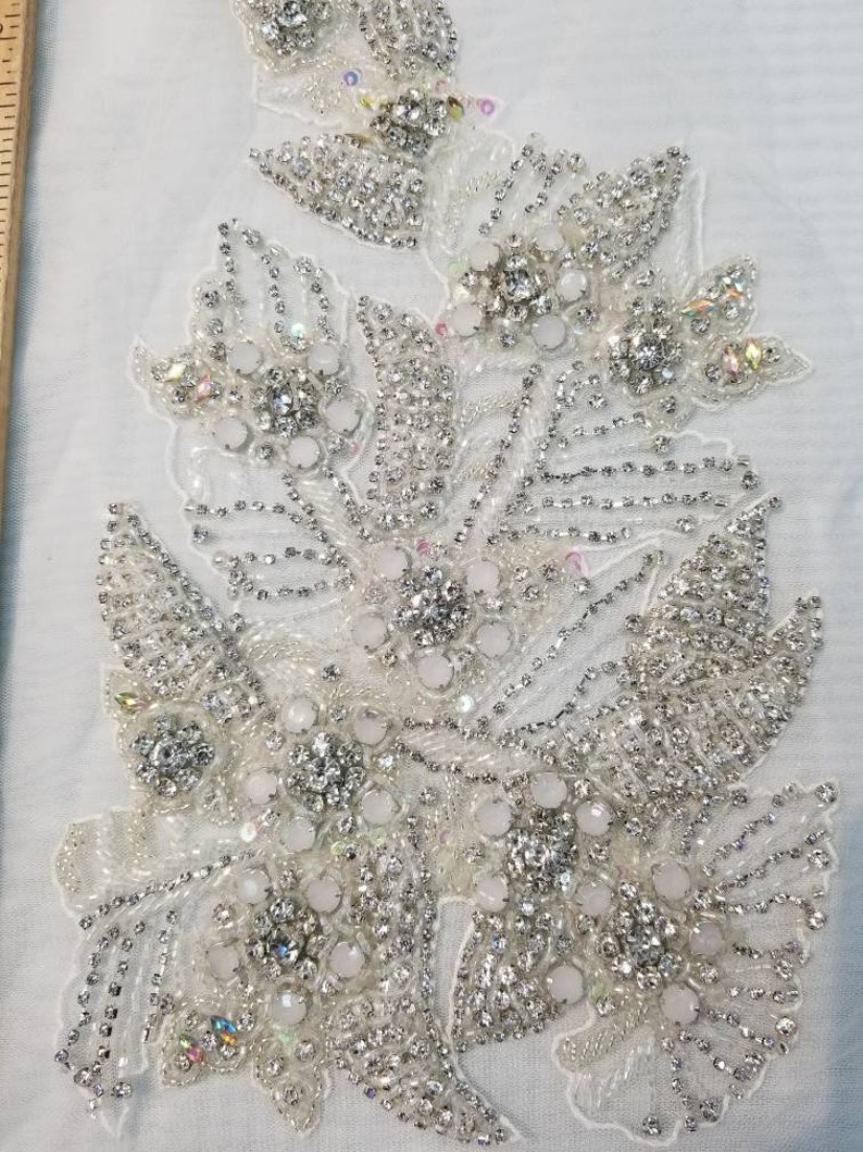 Peut inclure: Une applique florale blanche avec des perles et des strass &eacute;tincelants. Le motif pr&eacute;sente des grappes de fleurs et de feuilles, avec un m&eacute;lange d'ornements clairs et iris&eacute;s. L'applique est probablement destin&eacute;e &agrave; orner des v&ecirc;tements ou des accessoires.