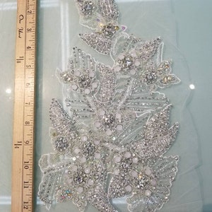 May include: A detailed, floral-shaped applique with silver beads, rhinestones, and white pearls. The applique is on a sheer, light blue fabric. The wooden ruler shows the applique is approximately 6 inches wide and 8 inches tall.