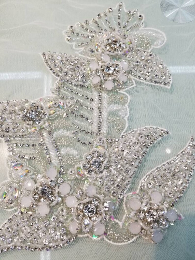 May include: A delicate floral design with white beads and clear rhinestones. The design is on a sheer, light green fabric.