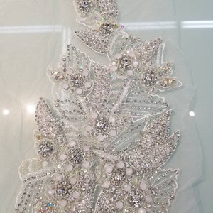 Peut inclure: Applique florale blanche d&eacute;taill&eacute;e sur un support transparent. Le motif pr&eacute;sente des grappes de perles blanches et de strass &eacute;tincelants, cr&eacute;ant un effet scintillant. L'applique est id&eacute;ale pour embellir les robes de mari&eacute;e ou autres v&ecirc;tements de c&eacute;r&eacute;monie.