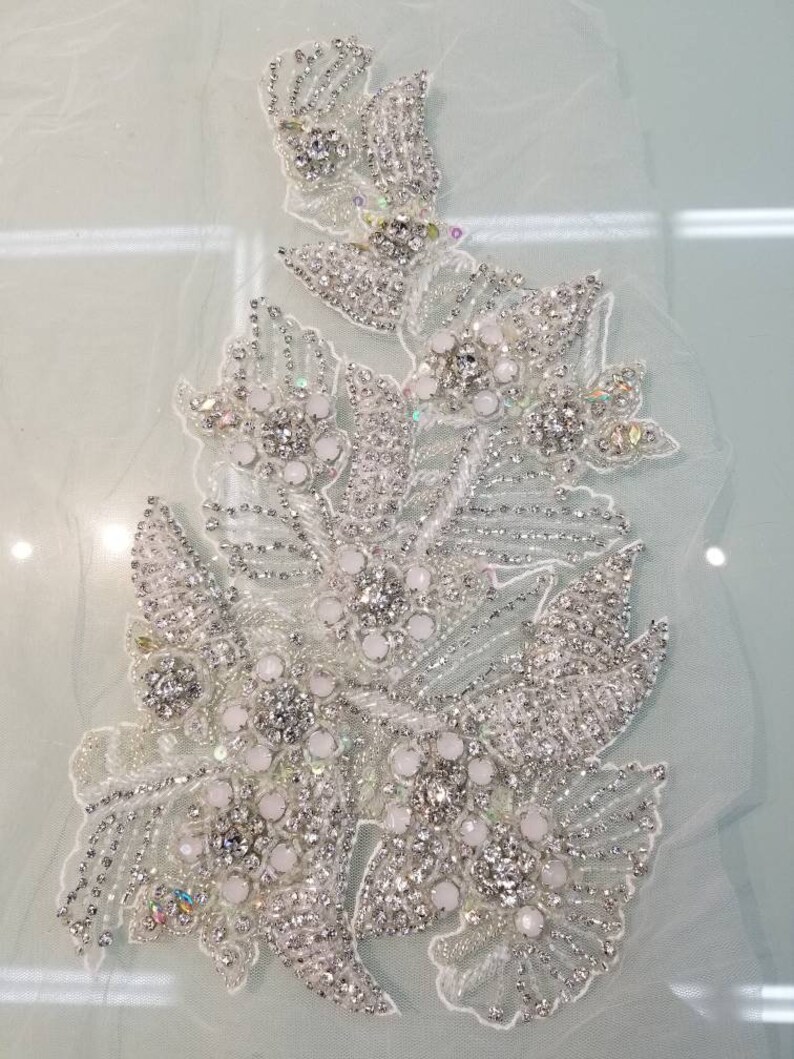 Peut inclure: Applique florale blanche d&eacute;taill&eacute;e sur un support transparent. Le motif pr&eacute;sente des grappes de perles blanches et de strass &eacute;tincelants, cr&eacute;ant un effet scintillant. L'applique est id&eacute;ale pour embellir les robes de mari&eacute;e ou autres v&ecirc;tements de c&eacute;r&eacute;monie.